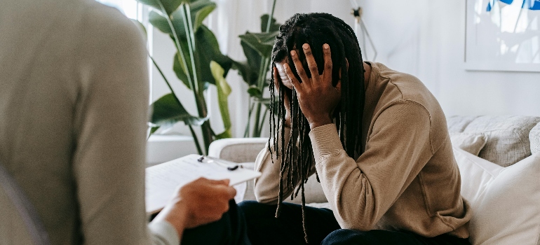 A person with dreadlocks sits on a couch, hands covering their face, while another person seated nearby holds a notebook. Green plants are visible.