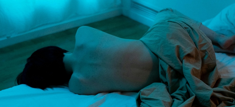 A person lies on a bed, their bare back exposed, partially covered by a light brown blanket in a softly lit, blue-toned room.