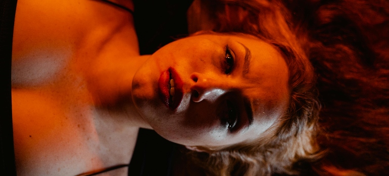 A close-up of a woman with red hair lying on her back, her expression intense, illuminated by warm, dramatic lighting.