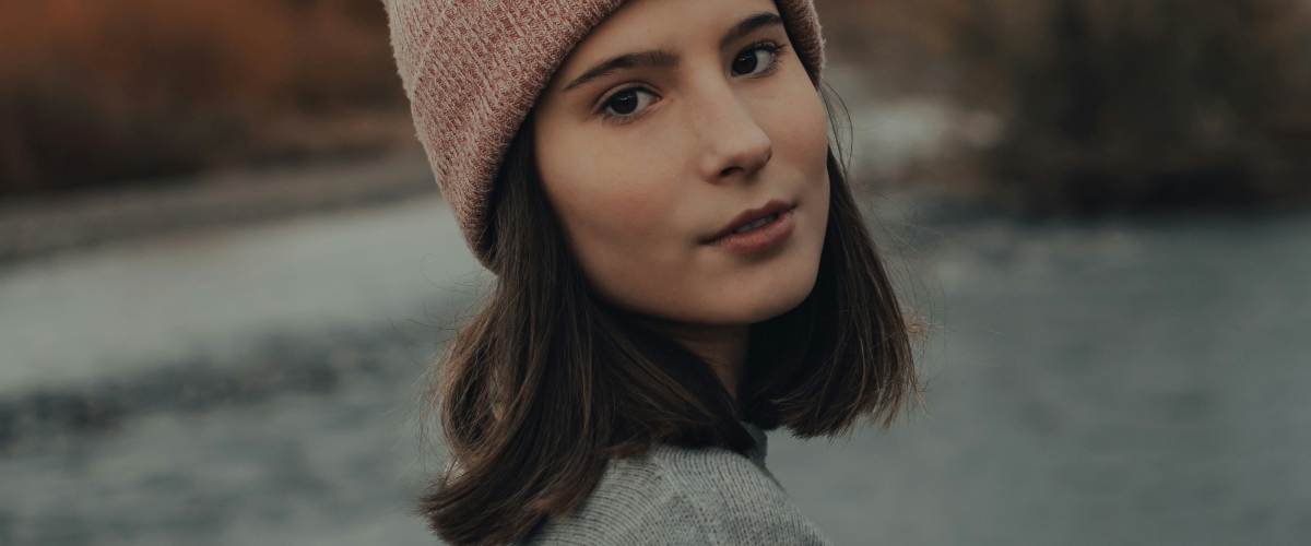 A woman wearing a pink knitted hat and a gray sweater stands by a tranquil body of water, surrounded by autumn foliage.