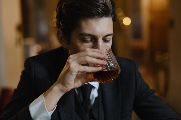A person in a suit holding a glass of amber-colored alcohol, symbolizing the link between PTSD and addiction.