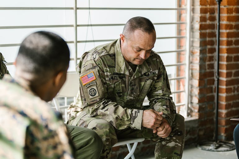 A military service member in camouflage sits in a sunlit room with fellow soldiers, reflecting the challenges of PTSD and addiction.