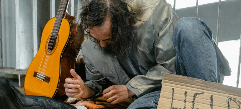 A person sits on the ground, holding a sign that says "HELP," next to a guitar and wearing a gray jacket.